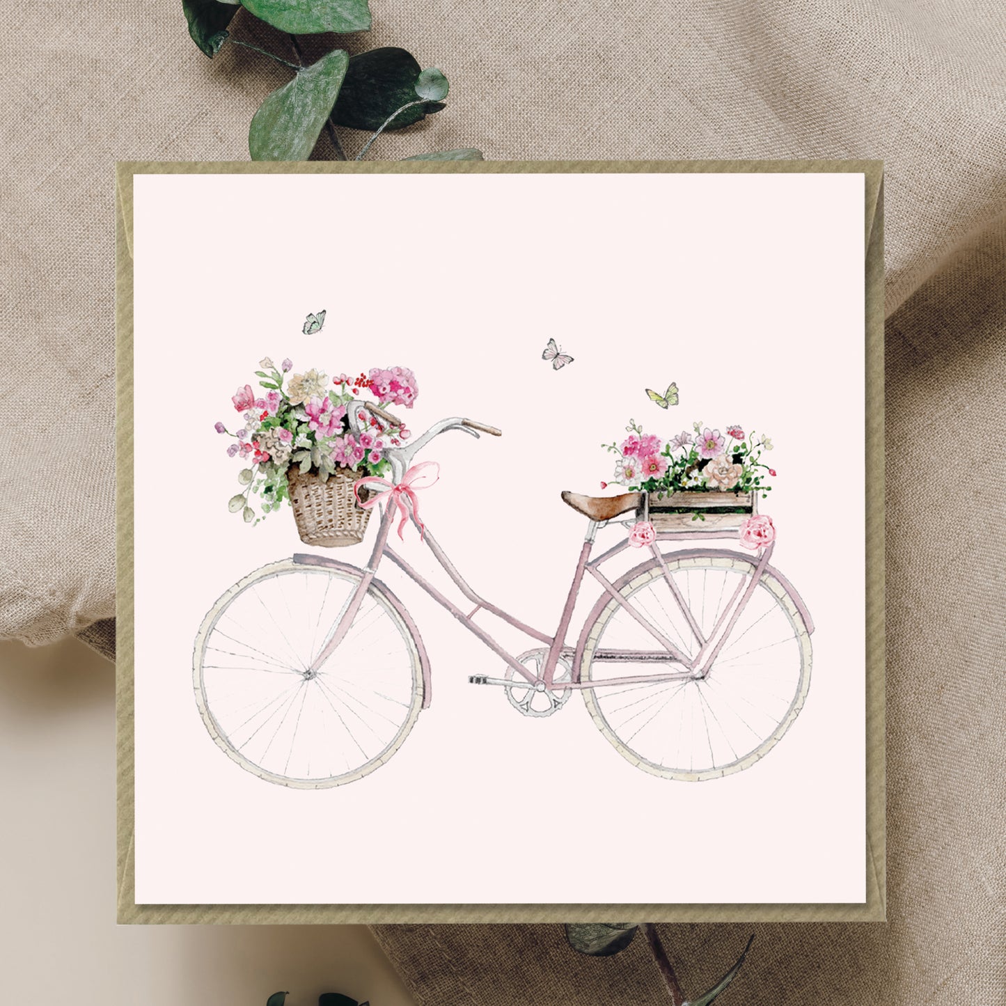 Greeting card with pastel bicycle, flower baskets, and butterflies on a neutral fabric background.