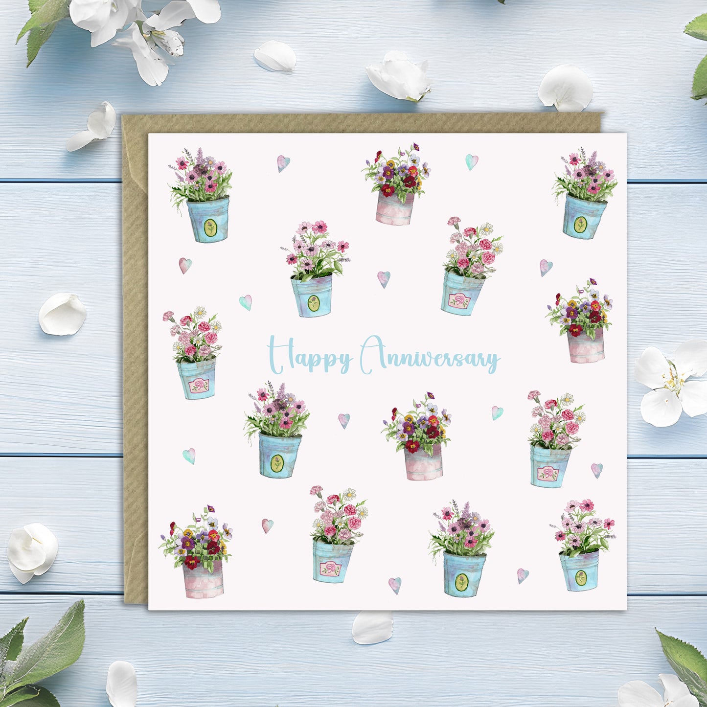 A square white anniversary greeting card with a kraft envelope, decorated with watercolor floral pots and the words 'Happy Anniversary' in light blue script. The card is photographed on a light blue wooden surface with scattered white flower petals around it.