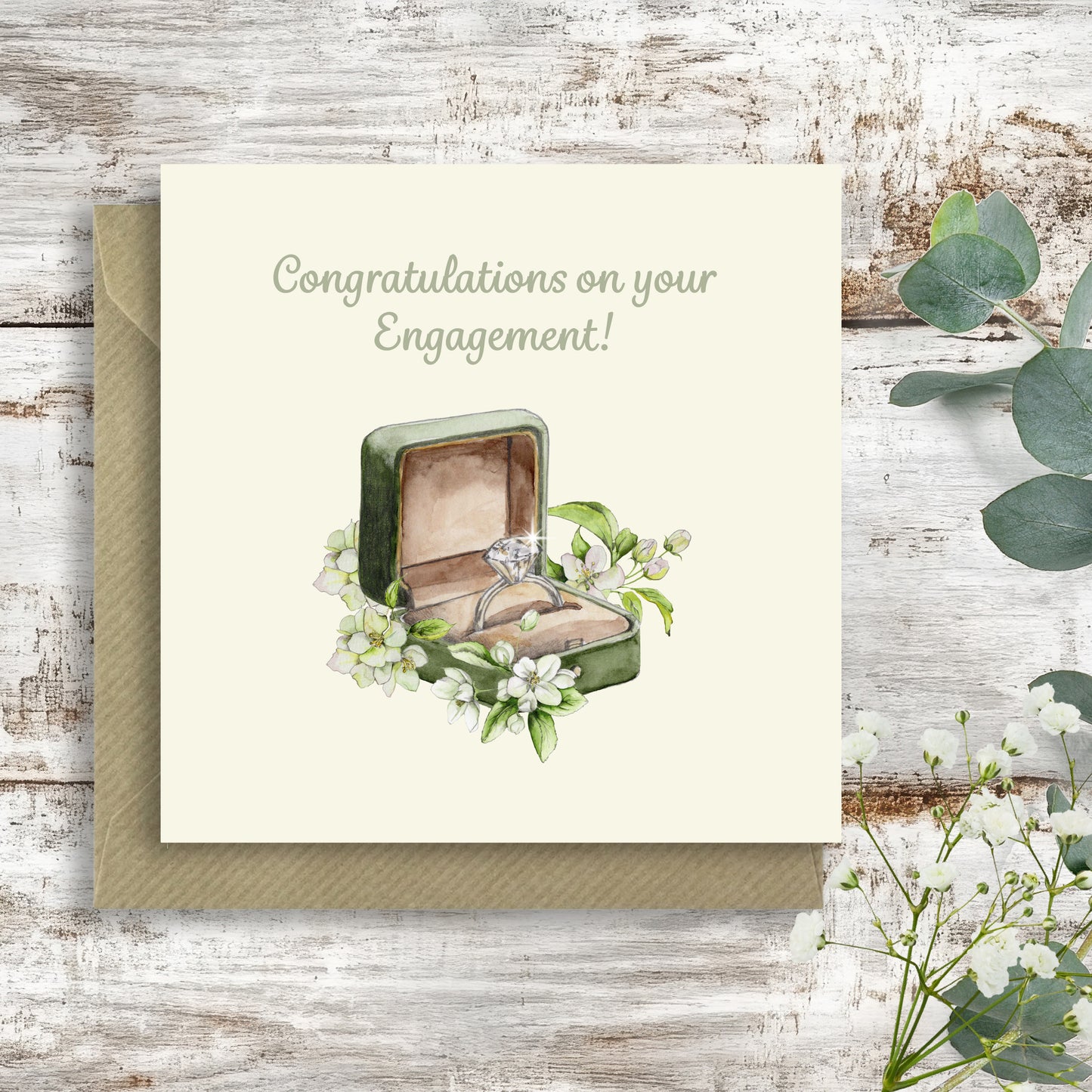 An engagement congratulations card with a matching envelope, featuring a watercolor illustration of an open ring box with a diamond ring and white flowers, displayed on a rustic white wood background with eucalyptus leaves.